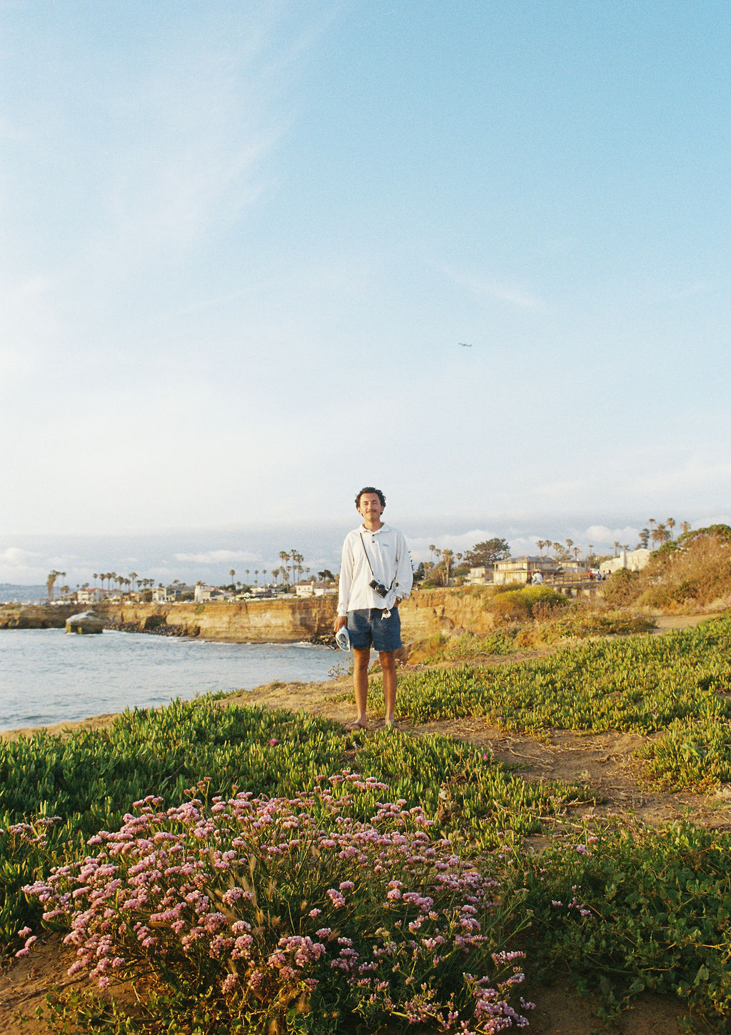 Man standing on grassy coastal bluff with pink flowers, ocean, and distant houses under a blue sky.