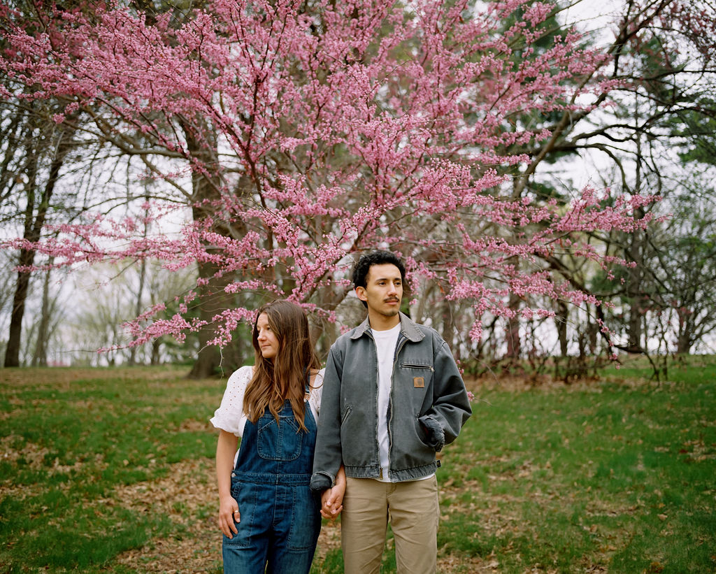 Couple holding hands standing outdoors in front of a tree with blooming pink flowers.