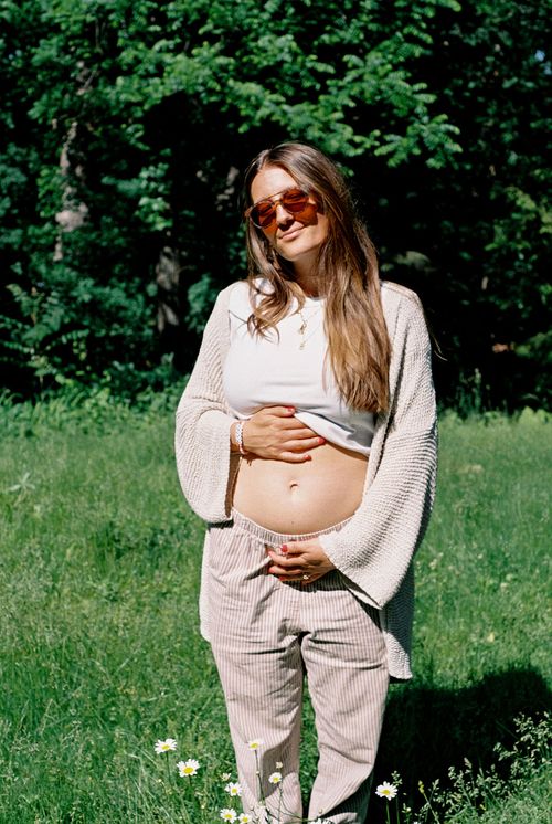 Pregnant woman in sunglasses standing outdoors with hands on her belly surrounded by grass and flowers.