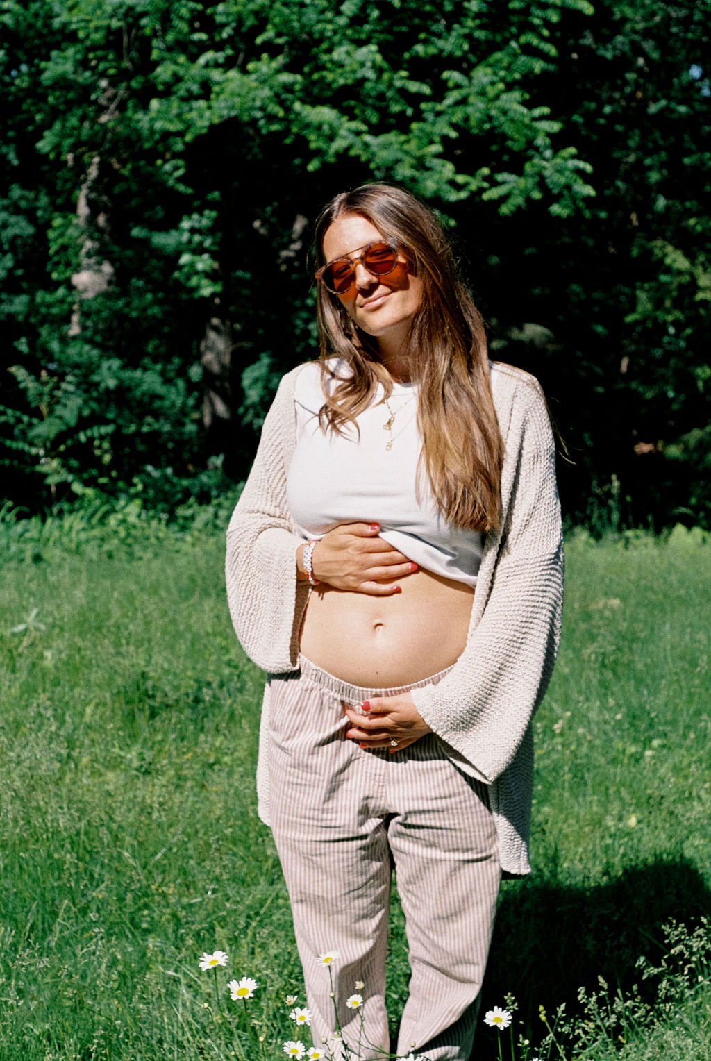 Pregnant woman in sunglasses standing outdoors with hands on her belly surrounded by grass and flowers.