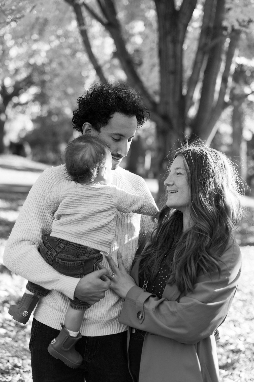 Man holding a baby while smiling woman touches the baby's arm outdoors in a park.