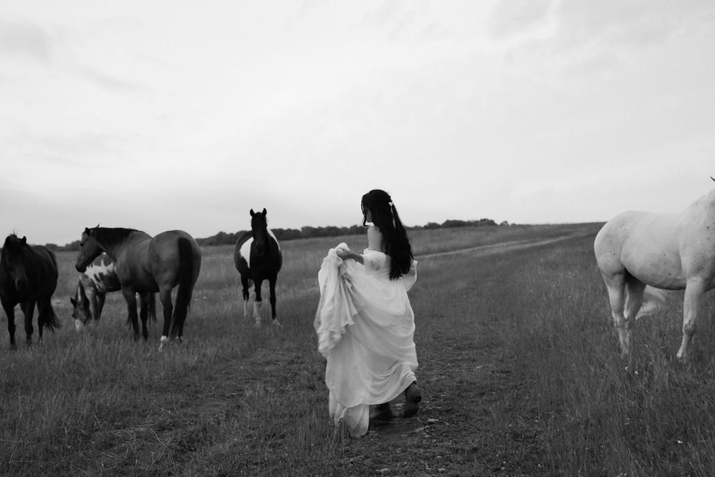 Woman in a flowing dress walking on a dirt path in a field surrounded by horses.