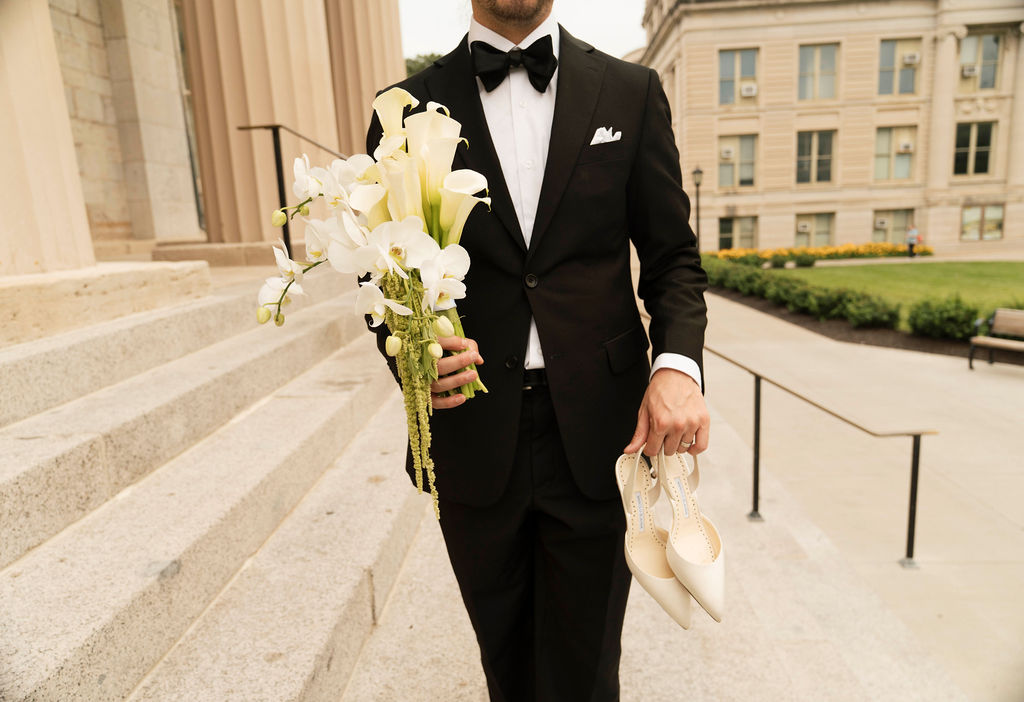 Man dressed in a black tuxedo holding a bouquet of white flowers in one hand and a pair of cream-colored high-heeled shoes in the other, standing on outdoor stone steps near a large building.