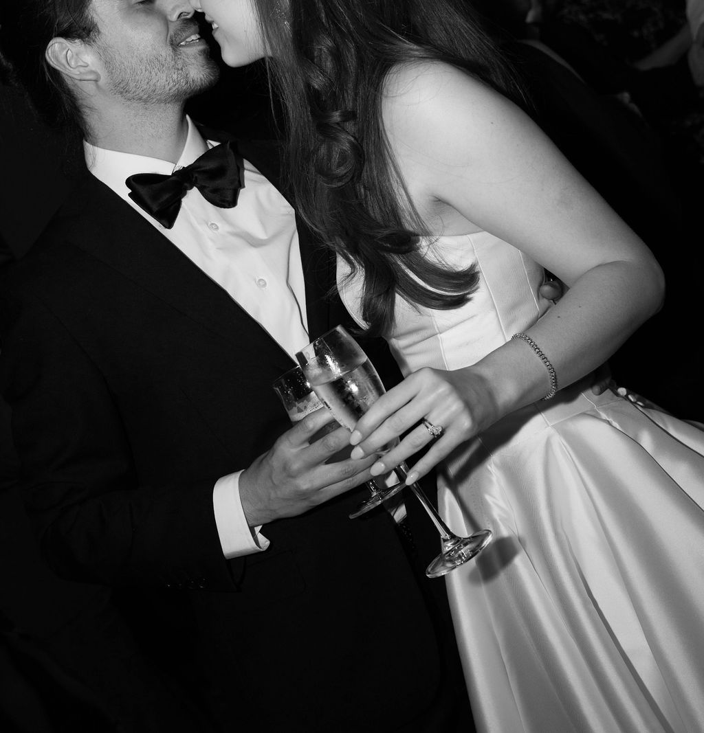 Black and white photo of a couple dressed formally, leaning in for a kiss while holding champagne glasses.
