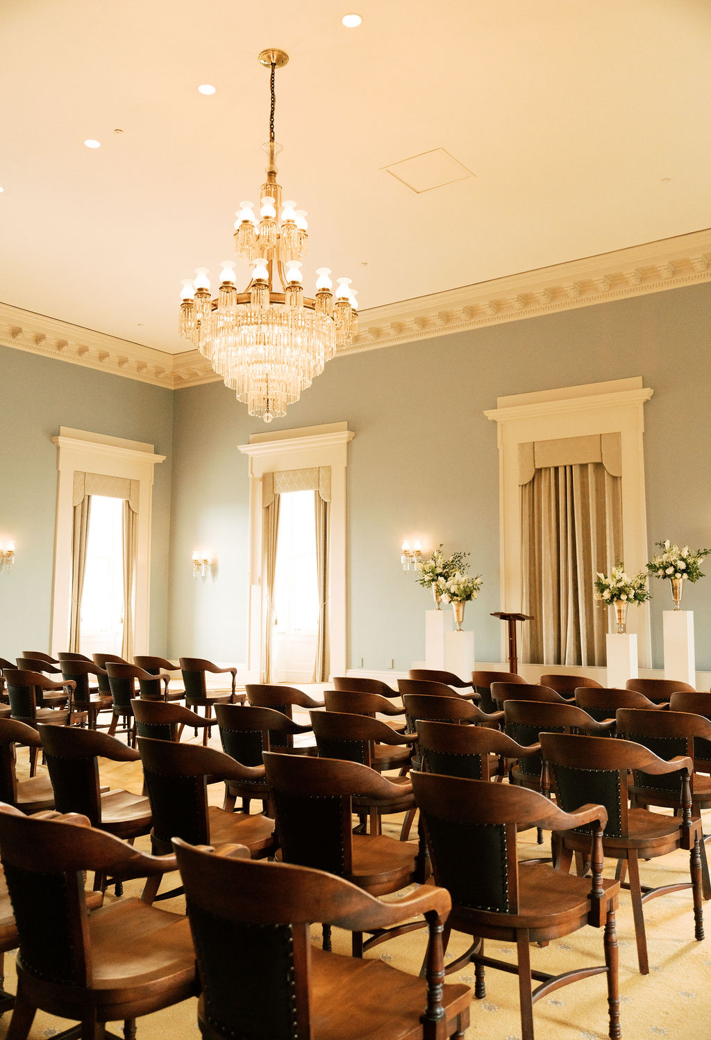 Elegant room with rows of wooden chairs, large chandelier, tall windows with curtains, and floral arrangements on pedestals.