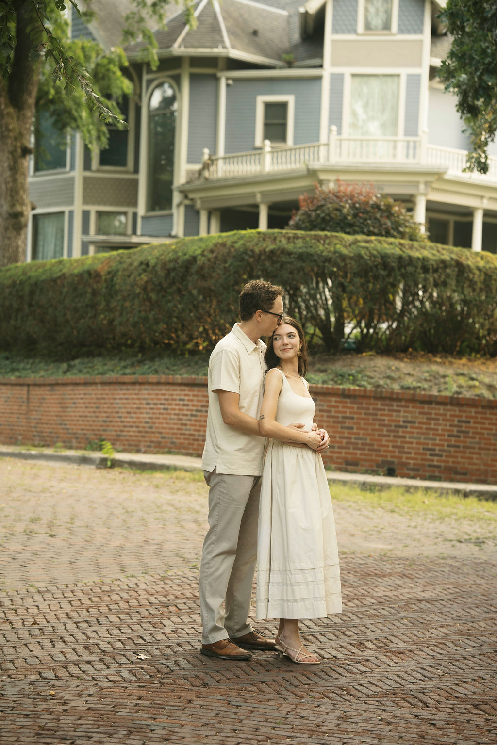 Couple embracing on a brick driveway in front of a large blue and gray house with white trim.