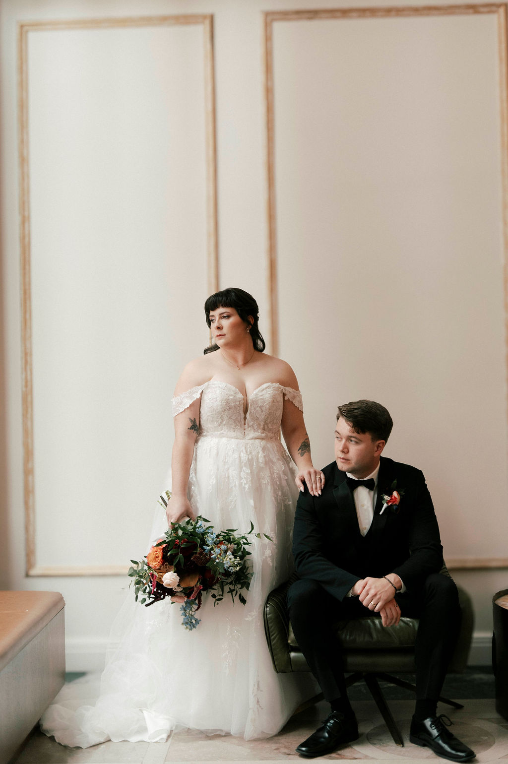 Bride in an off-shoulder white lace gown holding a colorful bouquet stands beside groom in a black tuxedo seated on a green chair in a softly lit room.