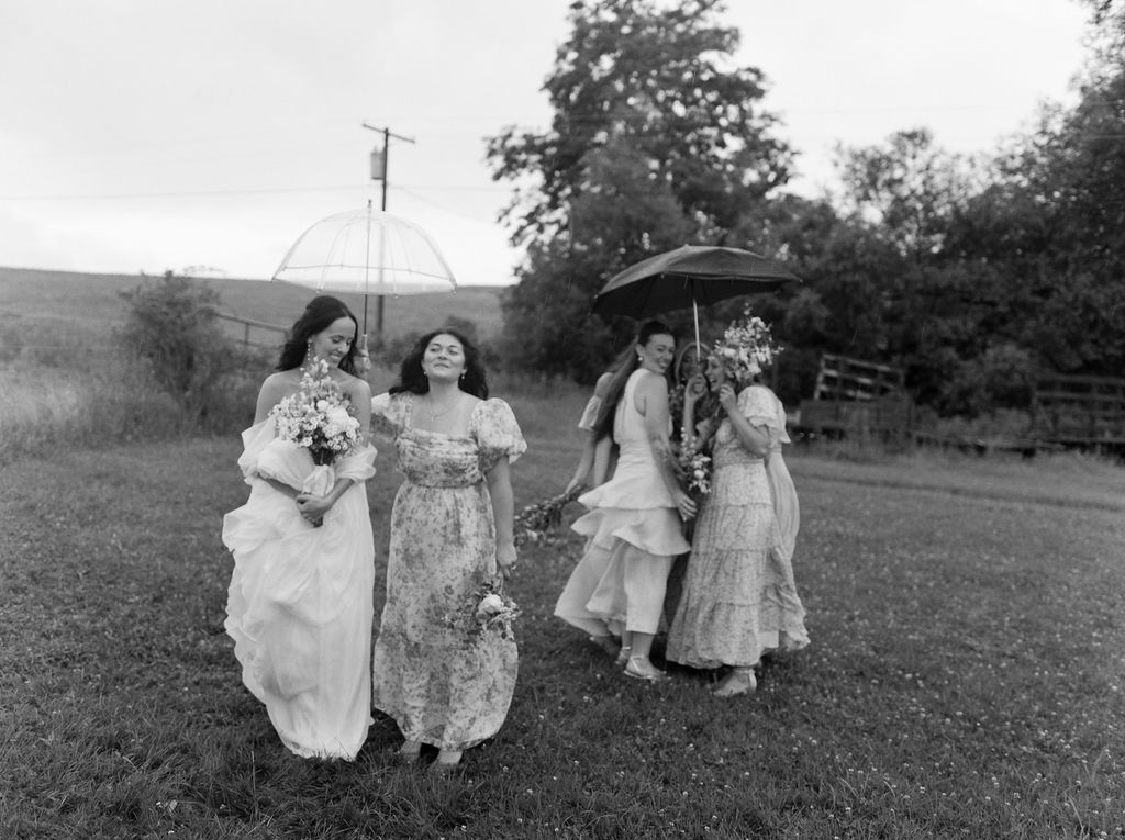 Group of women in dresses walking on grass, two holding umbrellas and flower bouquets in a rural outdoor setting.