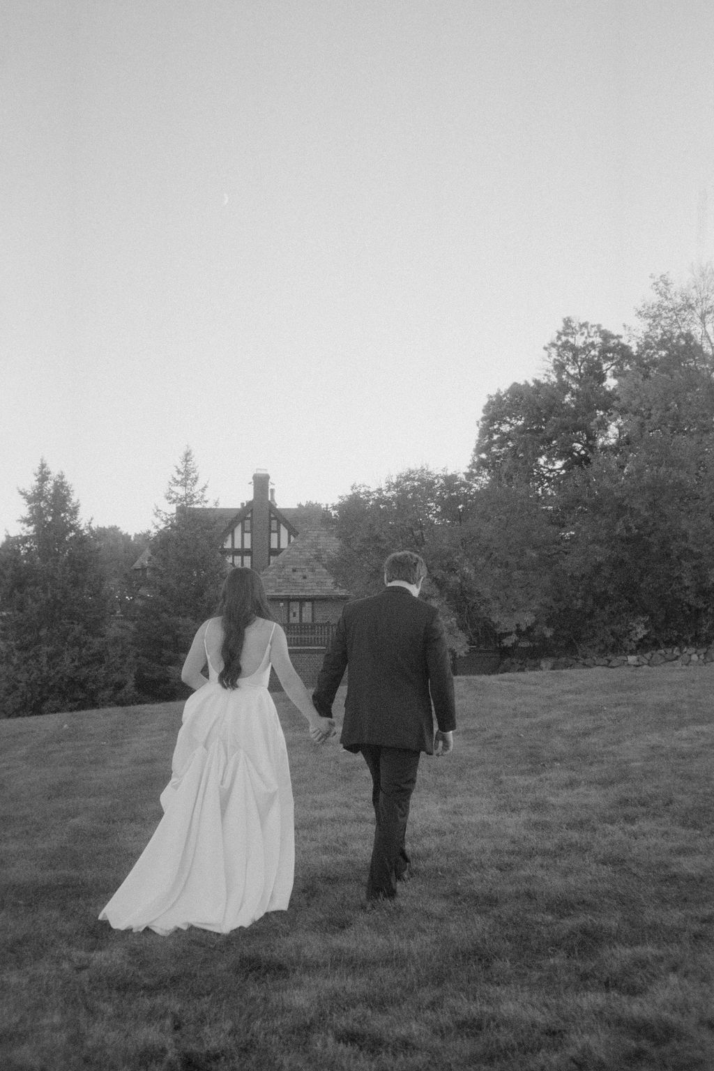 Bride and groom holding hands walking on grass toward a distant house surrounded by trees.