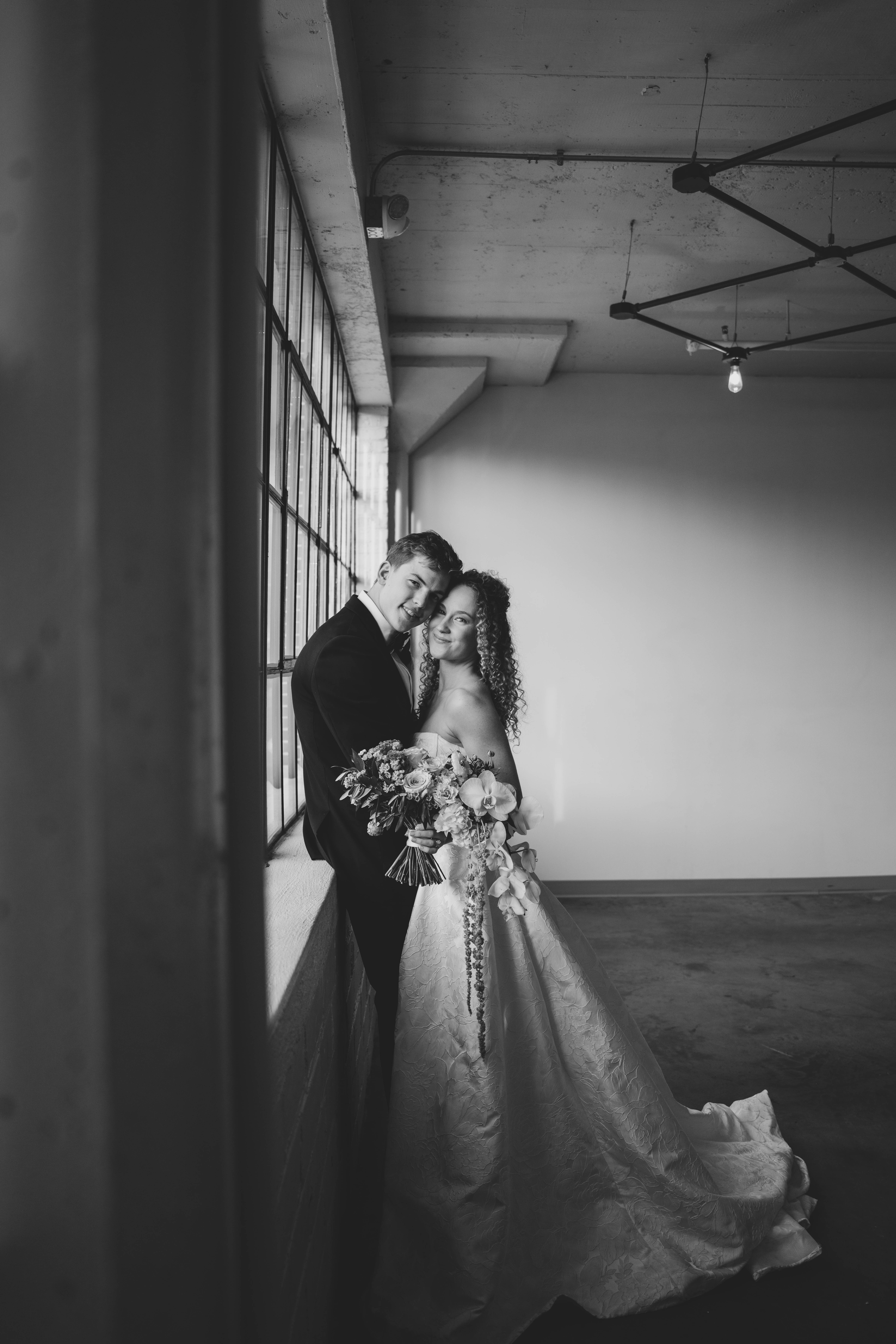 Black and white photo of a bride and groom gazing into each other's eyes in a softly lit room with a vintage wall lamp and shuttered window.