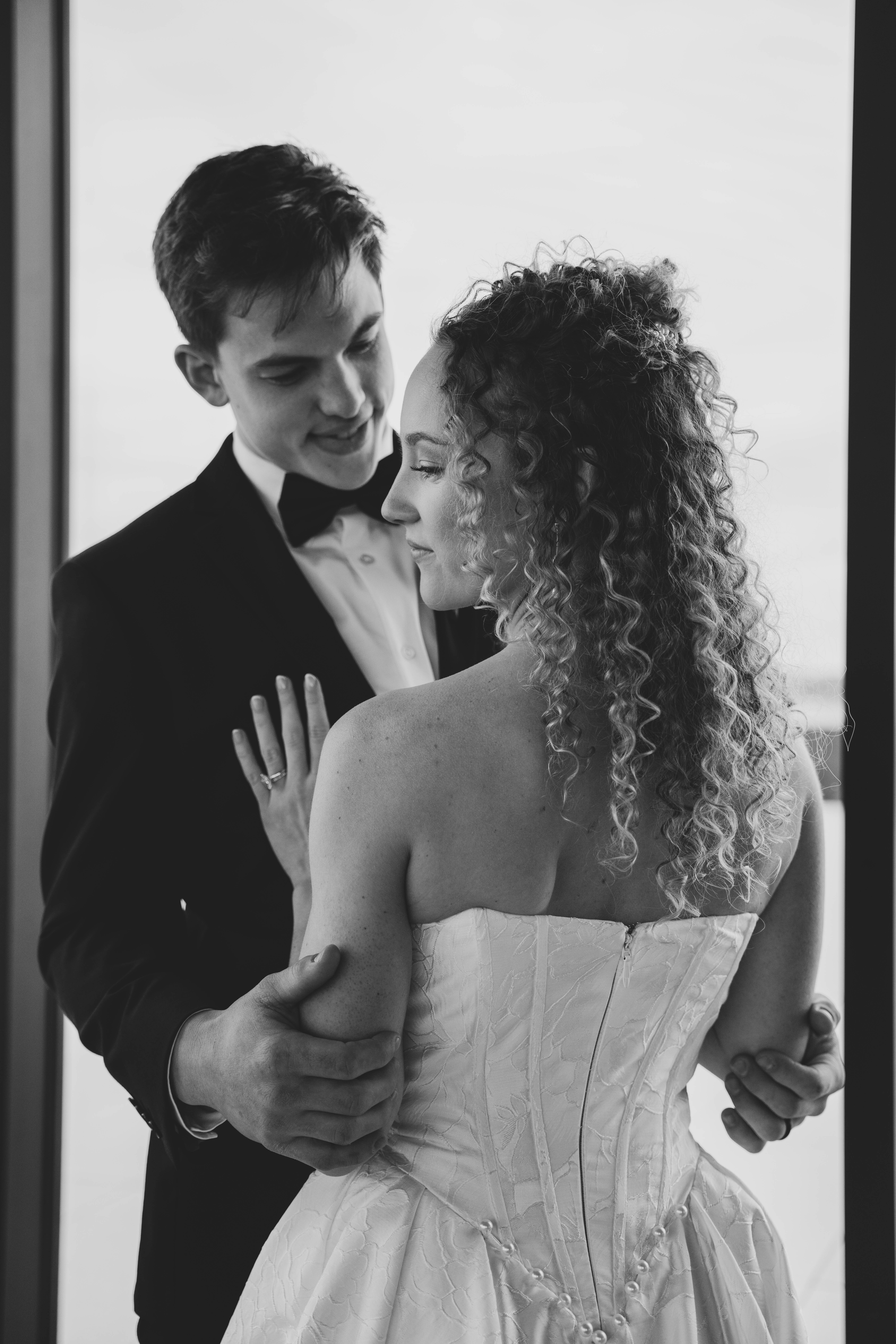 Black and white photo of a bride and groom embracing outdoors with a lake and trees in the background, the groom kissing the bride's temple under her veil.
