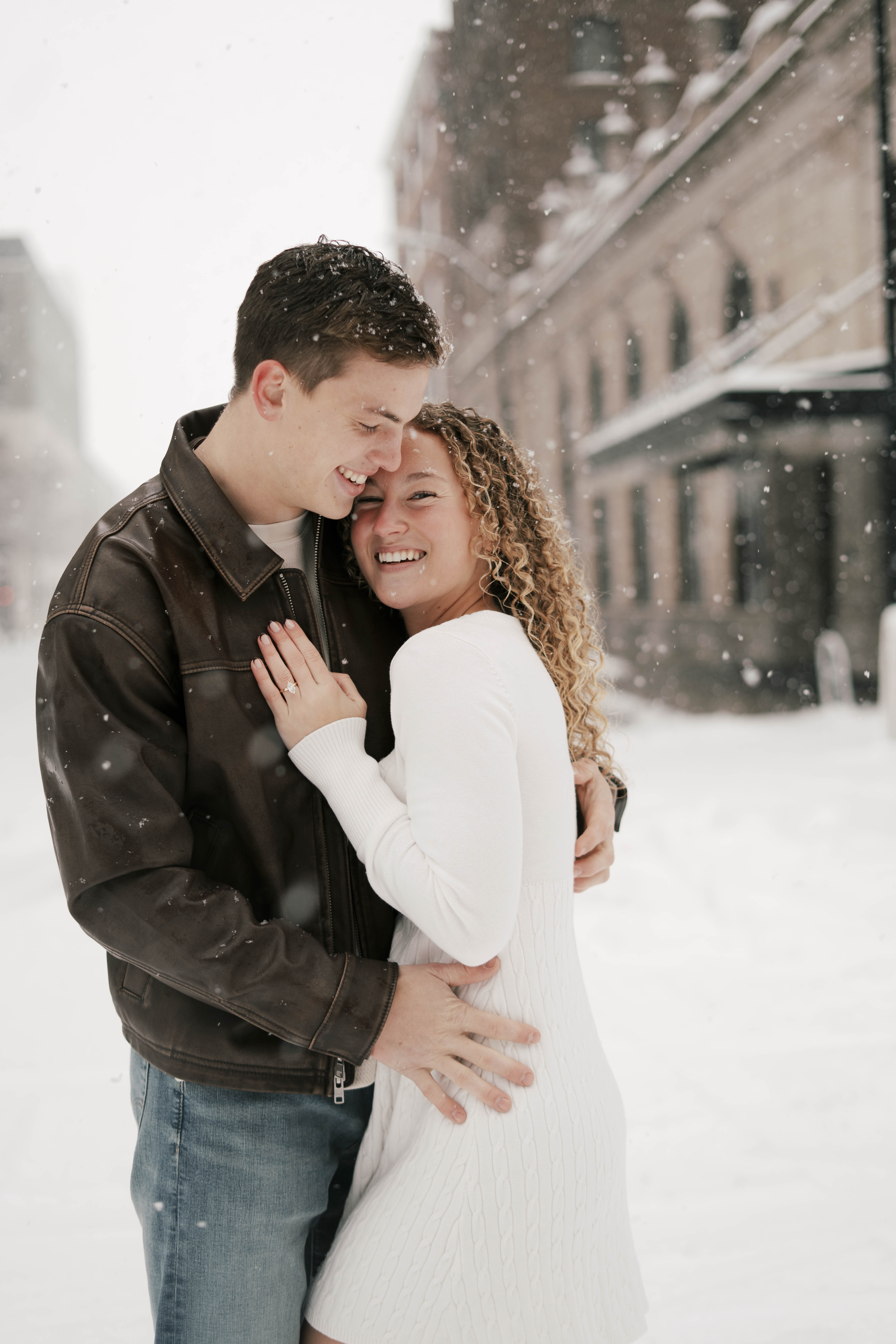 Bride in an off-shoulder white lace gown holding a colorful bouquet stands beside groom in a black tuxedo seated on a green chair in a softly lit room.