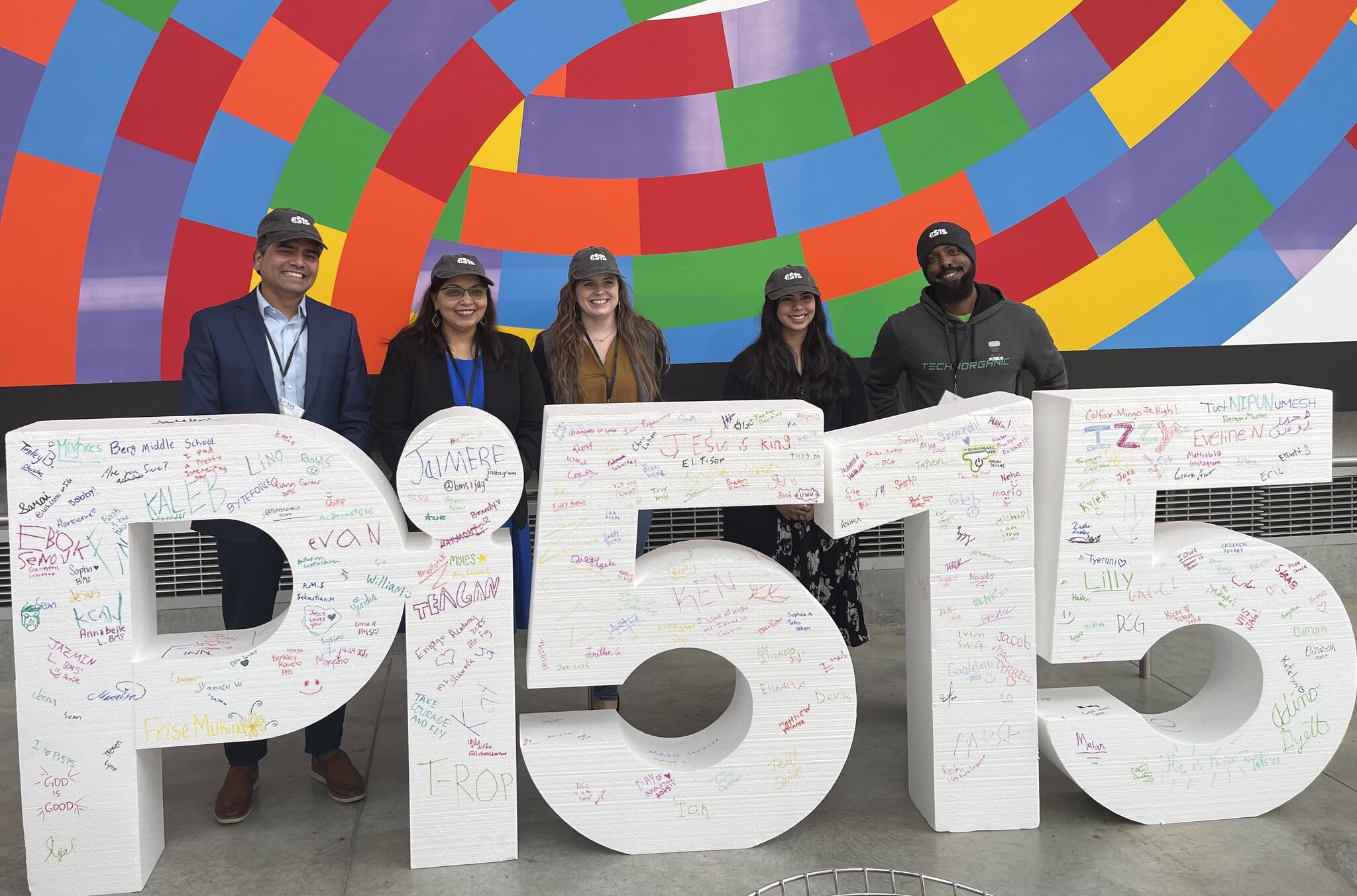 A busy cafe with people sitting at tables, chatting and using phones. Bright lighting, green pendant lamps, and shelves with colorful items create a lively atmosphere.