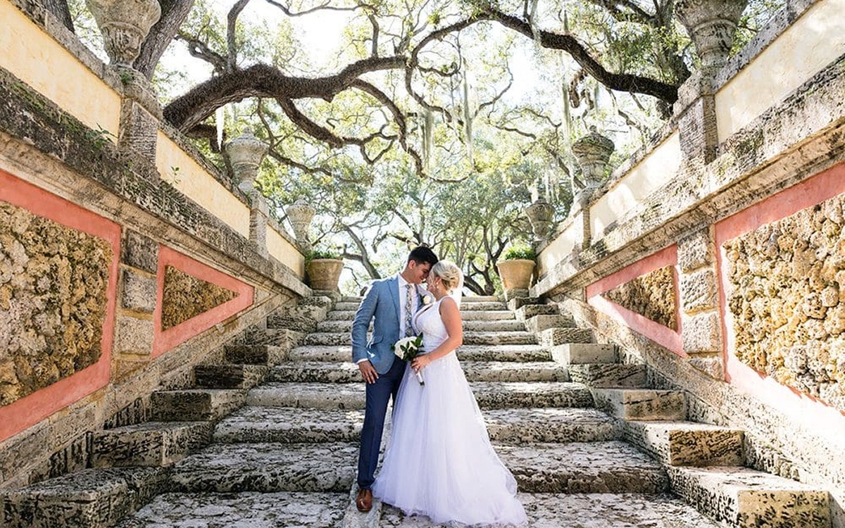 Bride and groom standing on a sandy beach with a lighthouse in the background, the bride holding a bouquet and a long veil flowing in the wind.