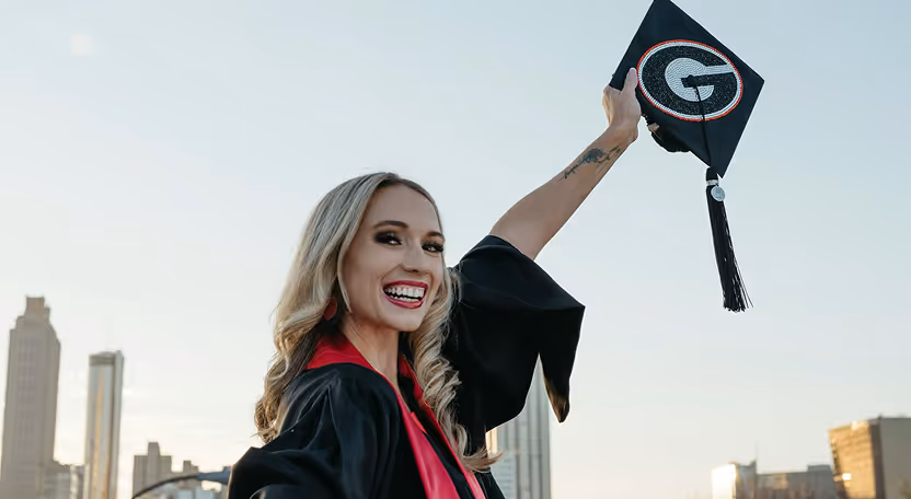 Smiling female graduate in black gown holding decorated graduation cap with a 'G' emblem against city skyline.