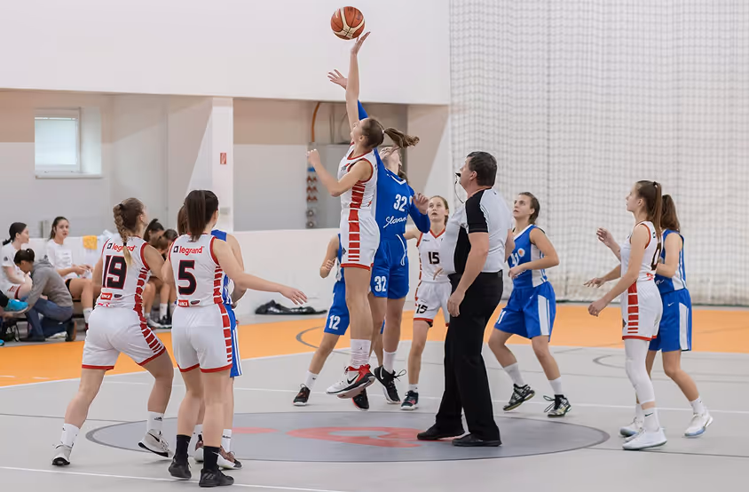 Girls' basketball game tip-off with players from blue and white-red teams jumping for the ball as the referee tosses it.