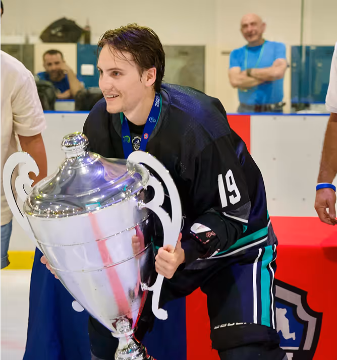 Young male ice hockey player wearing black jersey number 19 holding a large silver championship trophy on ice rink.