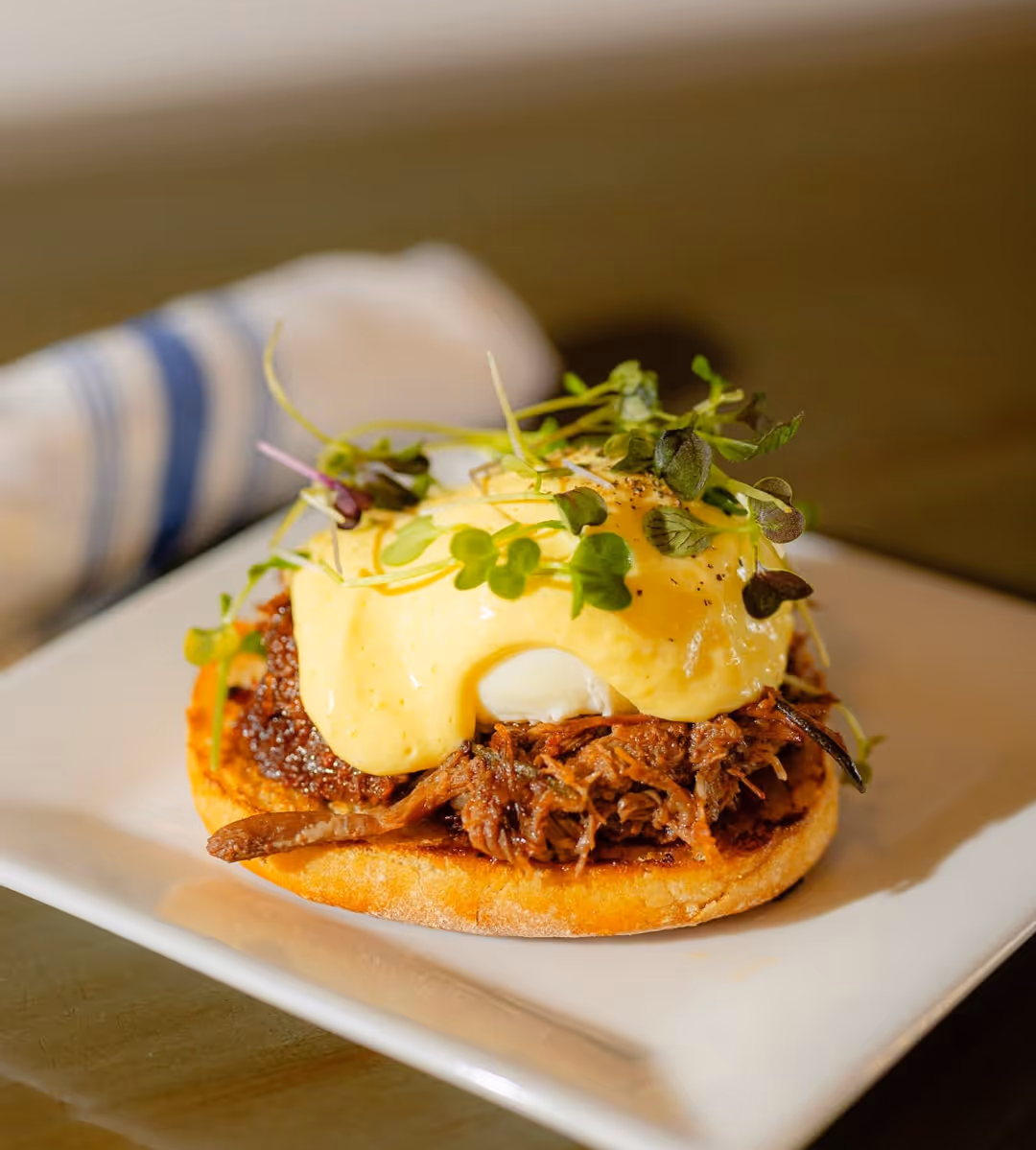 Eggs Benedict with pulled meat, hollandaise sauce, and microgreens on an English muffin.