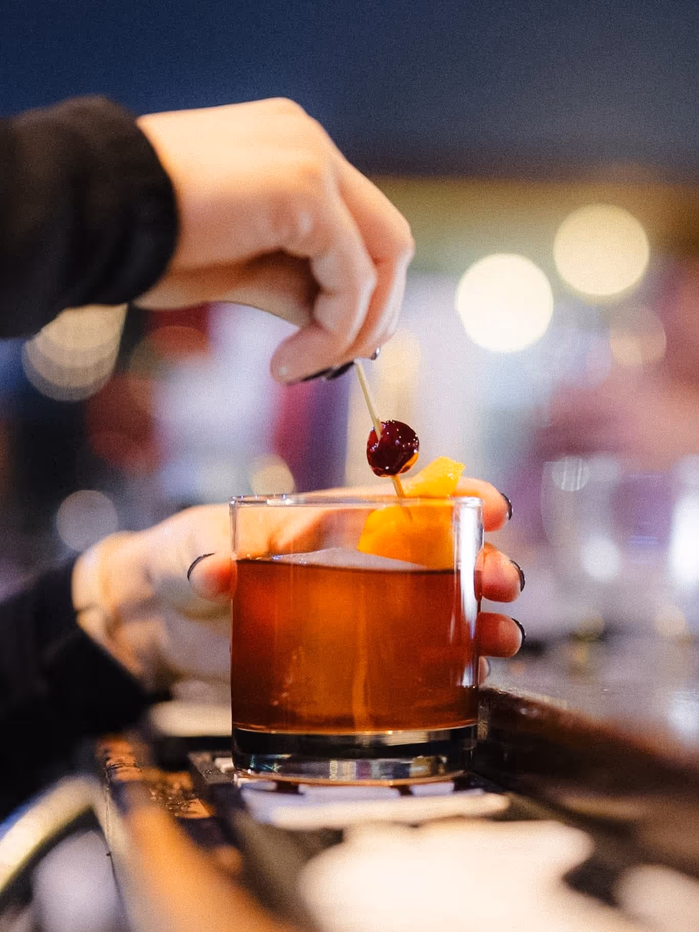 Hand garnishing an old fashioned with cherry and orange peel on bar counter with blurred lights.