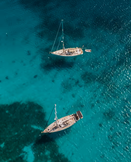 Aerial view of two sailboats anchored on clear turquoise water with visible underwater shadows.