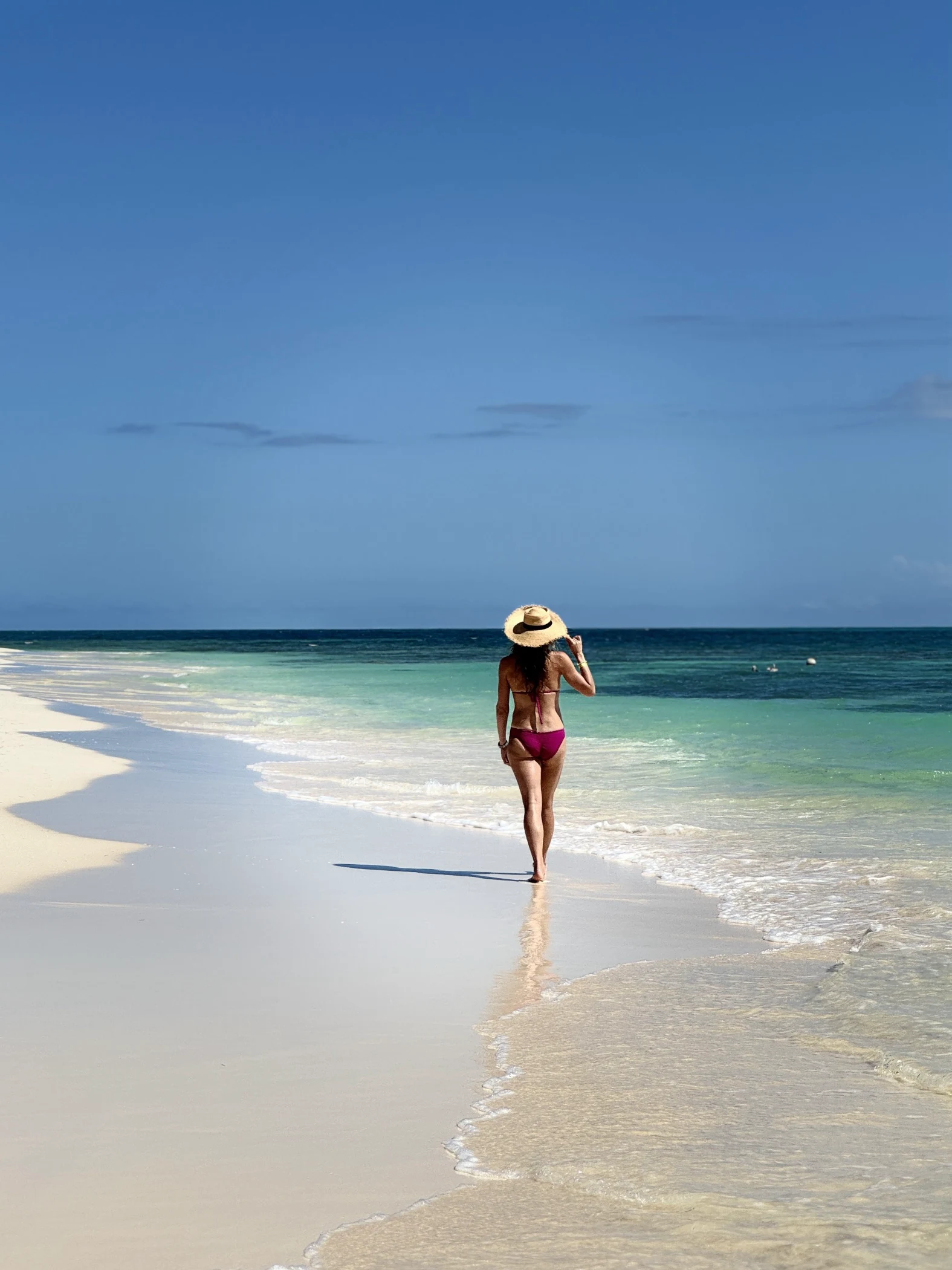 Woman in a pink bikini and sun hat walking on a sandy beach by clear turquoise ocean under blue sky.