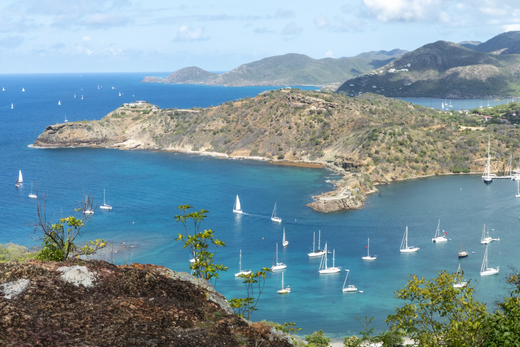 Coastal bay with numerous sailboats anchored near rocky, shrub-covered hills under a partly cloudy sky.