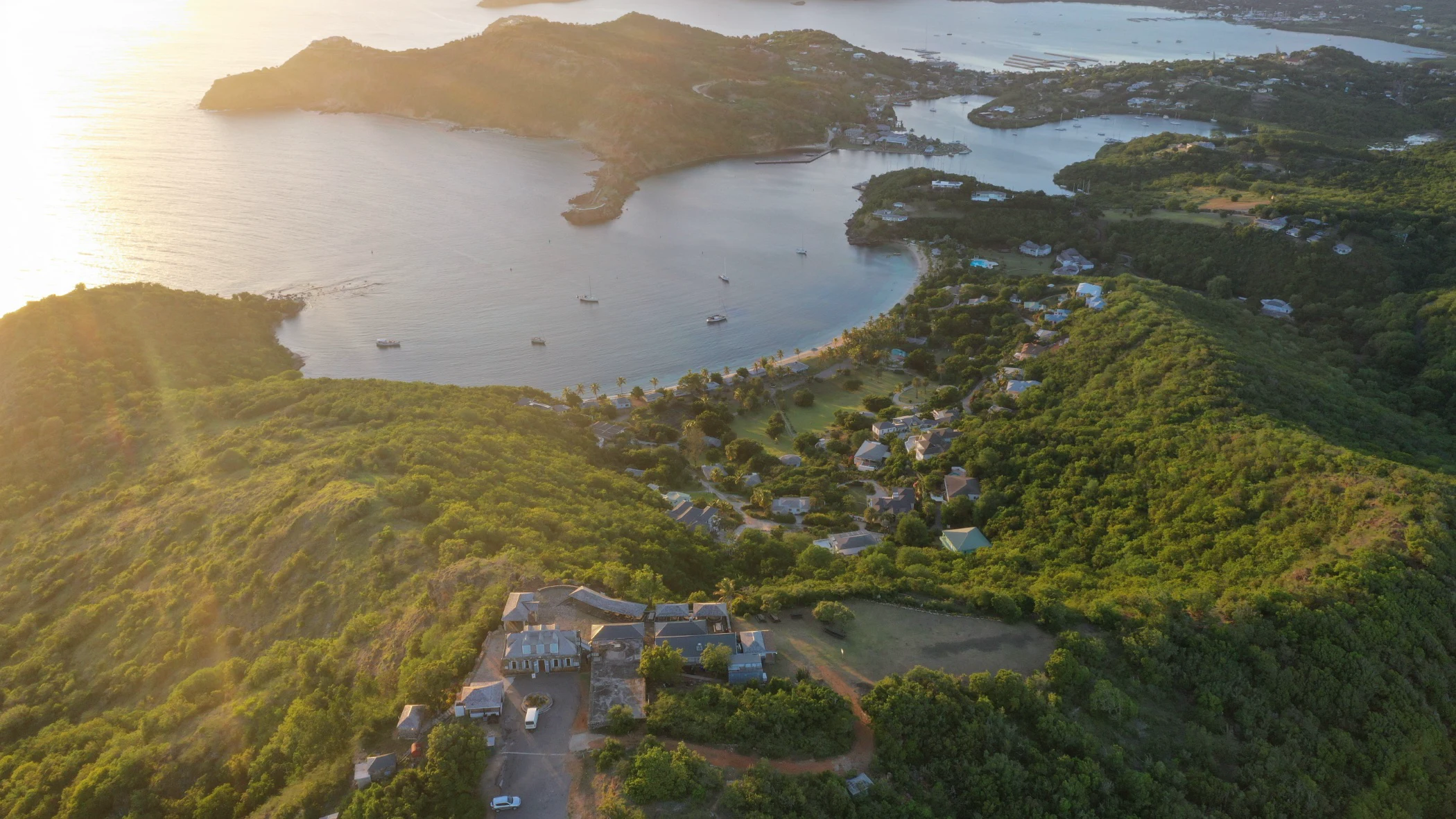 Aerial view of a coastal bay at sunset with boats anchored in calm water and houses nestled among green hills.