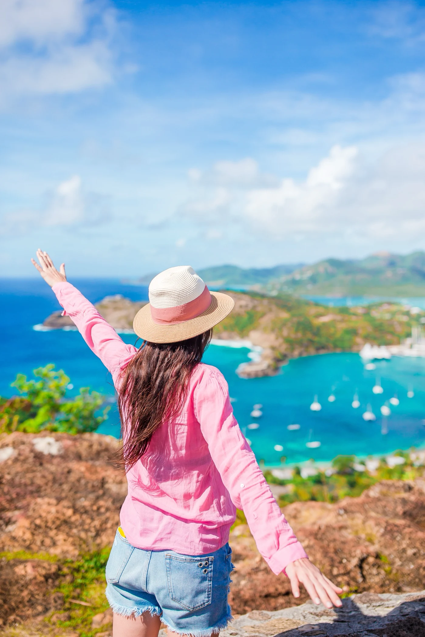 Woman in a pink shirt and straw hat with arms outstretched overlooking a blue bay with sailboats and green hills.