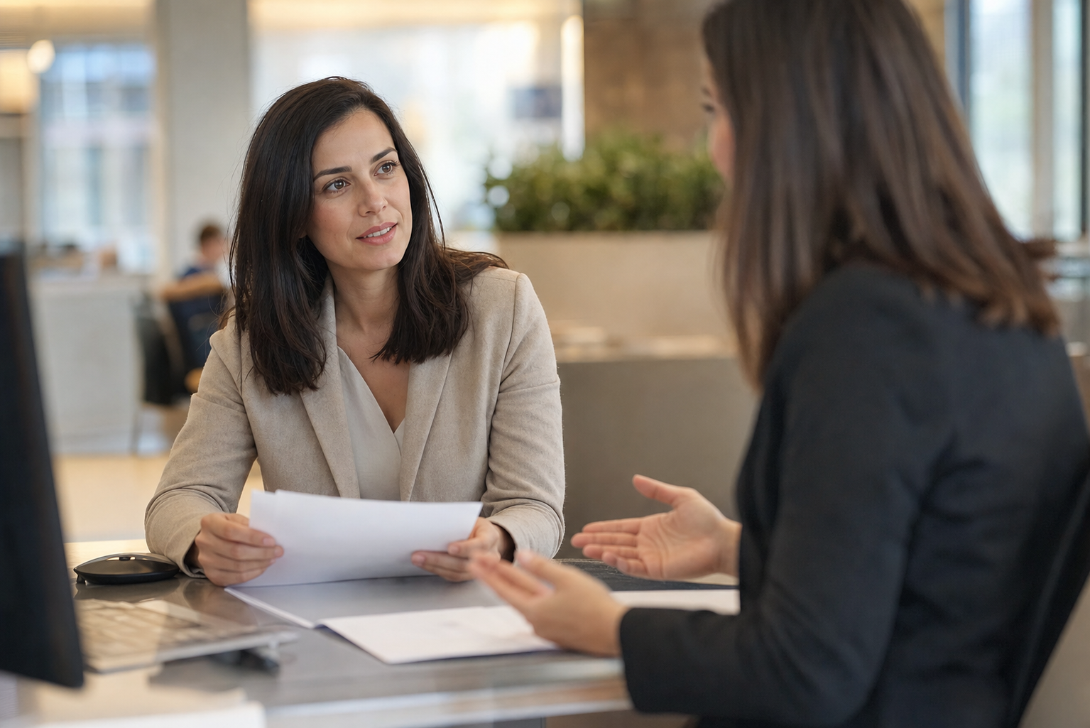 A photojournalistic photo of a professional woman sitting at a bank branch desk across from a bank representative, viewed from a natural side angle. The woman is mid-conversation, holding official documents in her hands. The setting is a modern bank office with warm natural lighting from windows. The bank representative's hands are visible gesturing toward paperwork on the desk. Shallow depth of field, embedded documentary style photography. Warm, real-life atmosphere. No text, words, or letters visible in the image.