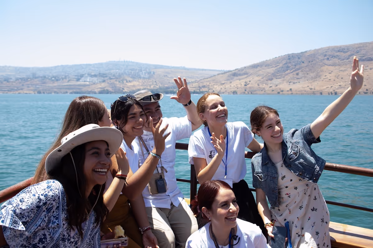 Grupo de seis jóvenes sonrientes posando y saludando mientras están en un bote con agua y colinas al fondo.