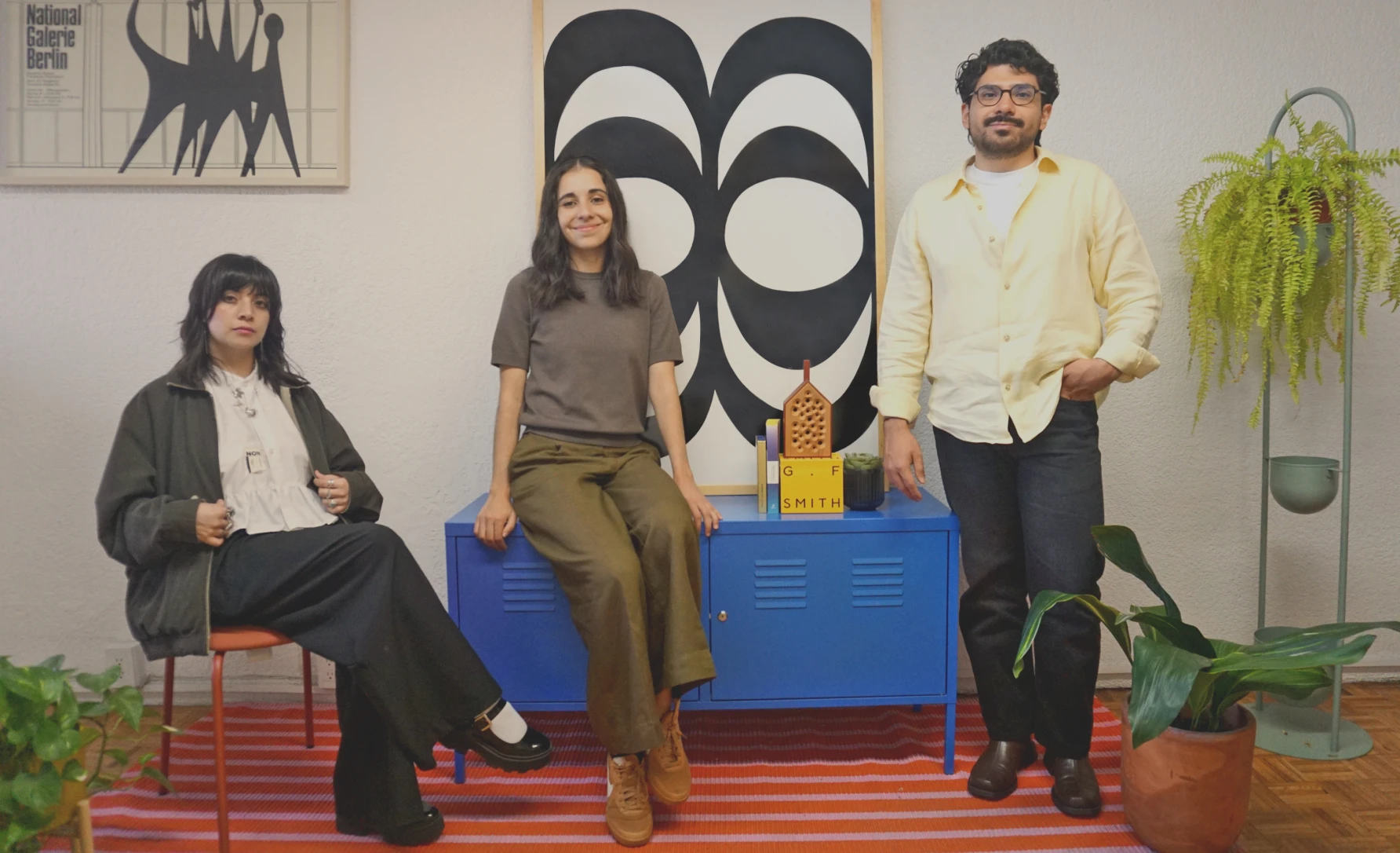 Three people posing indoors: one seated with crossed legs, one sitting on a blue cabinet, one standing beside it.