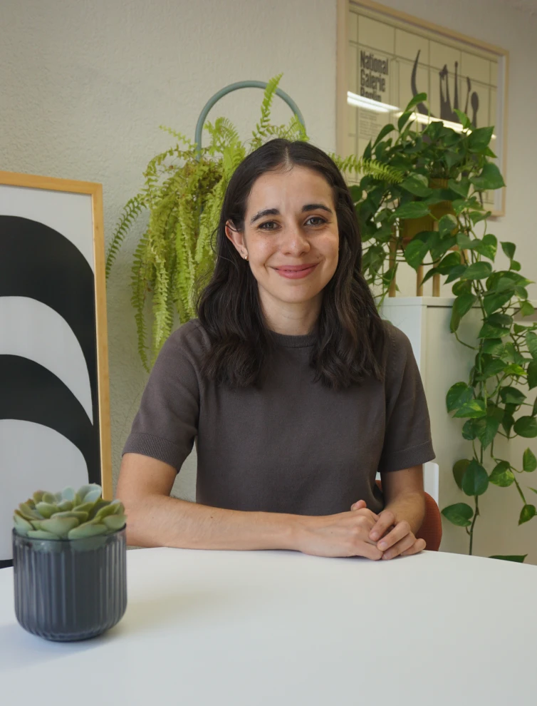 Woman with dark hair and brown shirt sitting at a white table with green plants behind her.