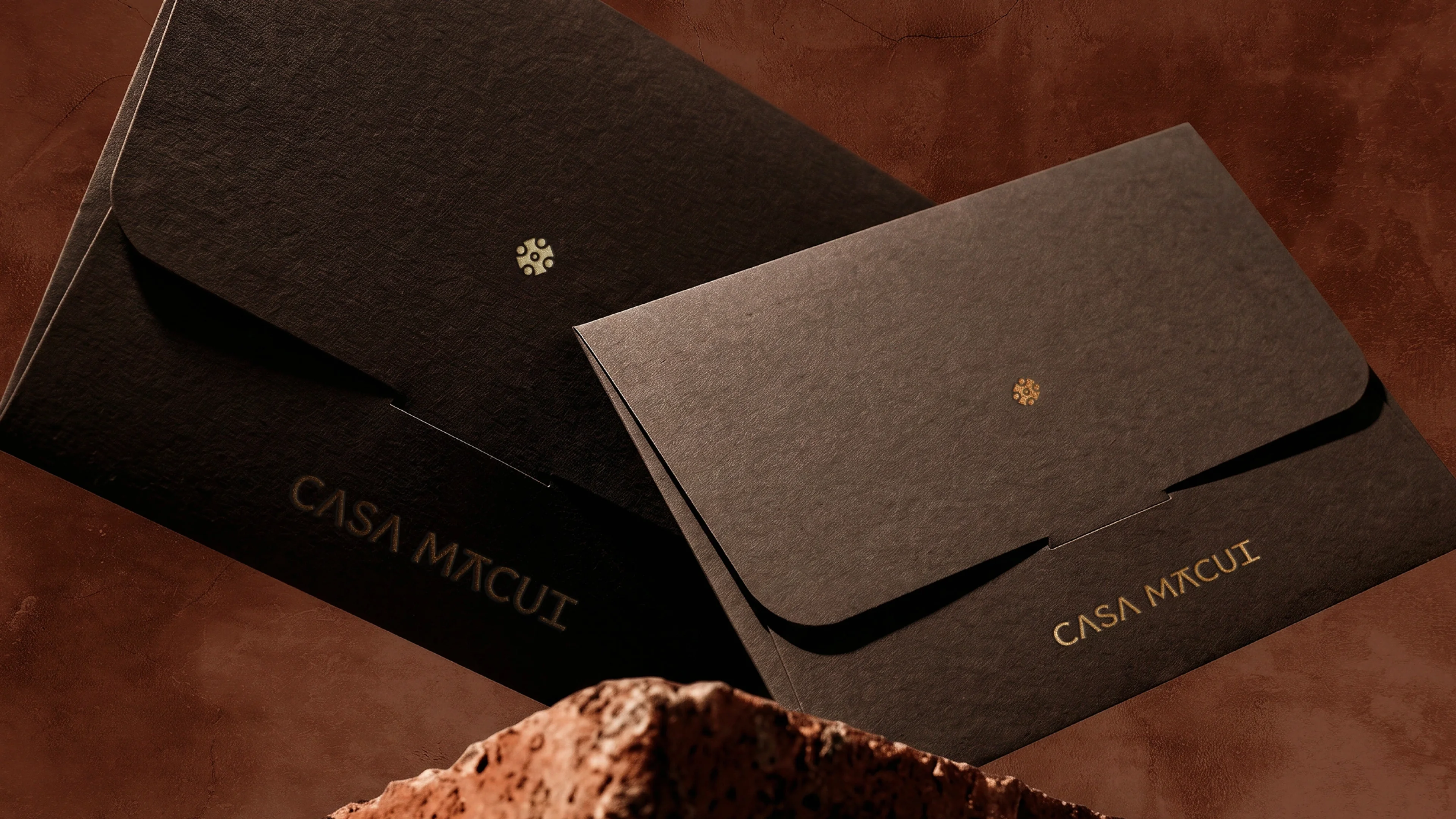 Two dark brown branded envelopes balanced on a rough rock against a terracotta background.