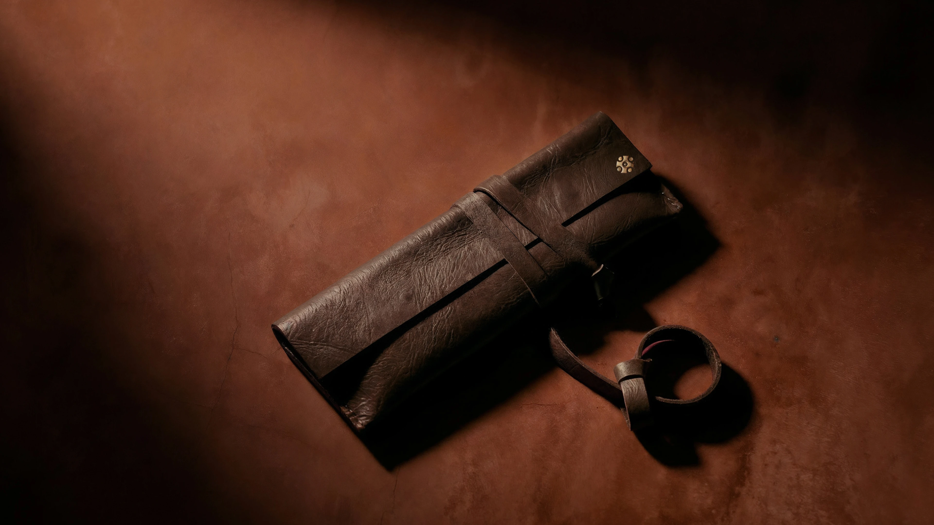 A dark brown leather knife roll on a textured terracotta surface under dramatic lighting.