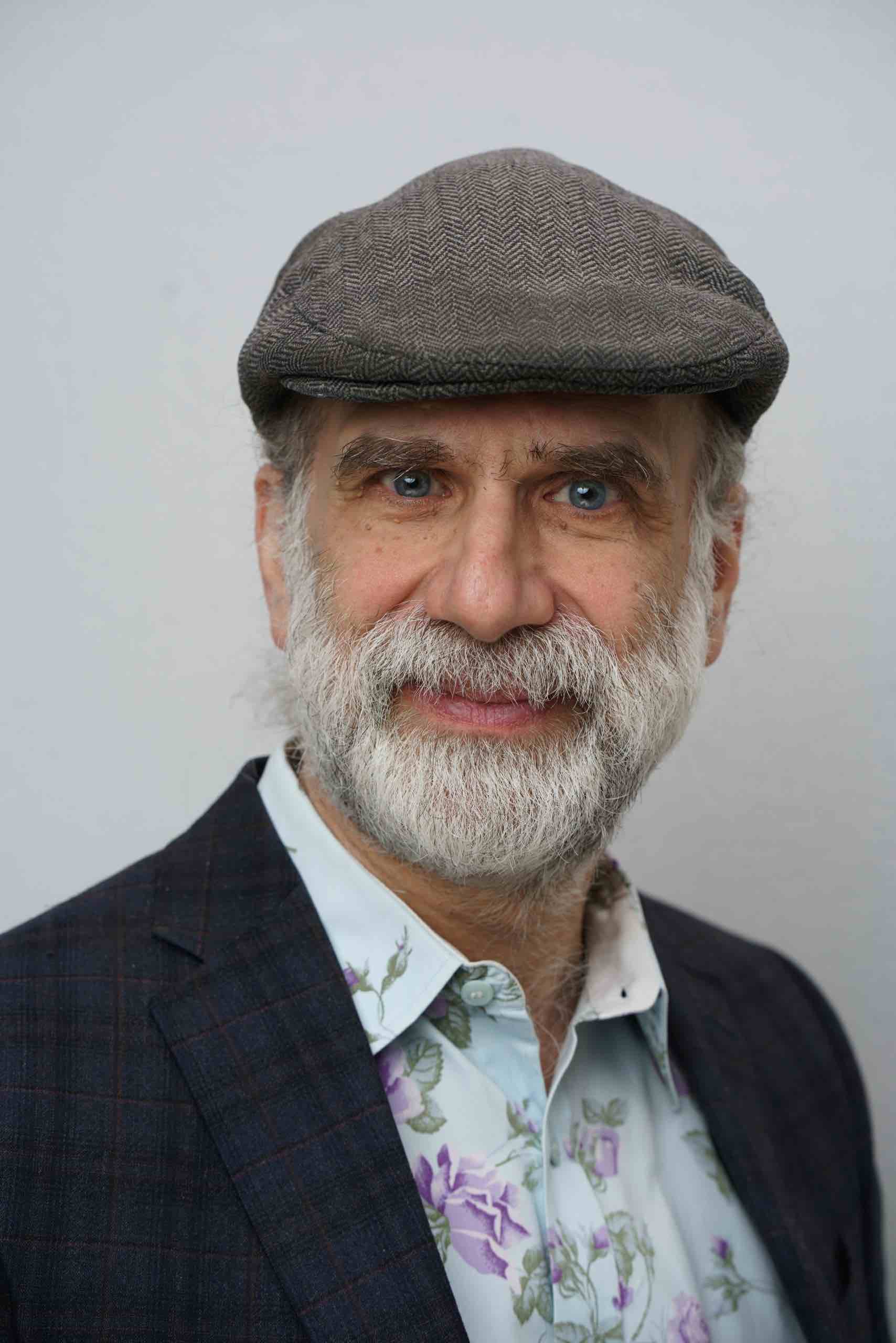 Older man with white beard wearing a gray flat cap, floral shirt, and dark plaid jacket smiling slightly.