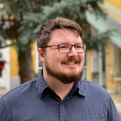 Smiling man with glasses, beard, and mustache wearing a blue button-up shirt standing outdoors near a tree and yellow building.