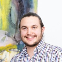 Smiling man with light skin and long dark hair tied back, wearing a checkered shirt, standing indoors against a colorful abstract painting.
