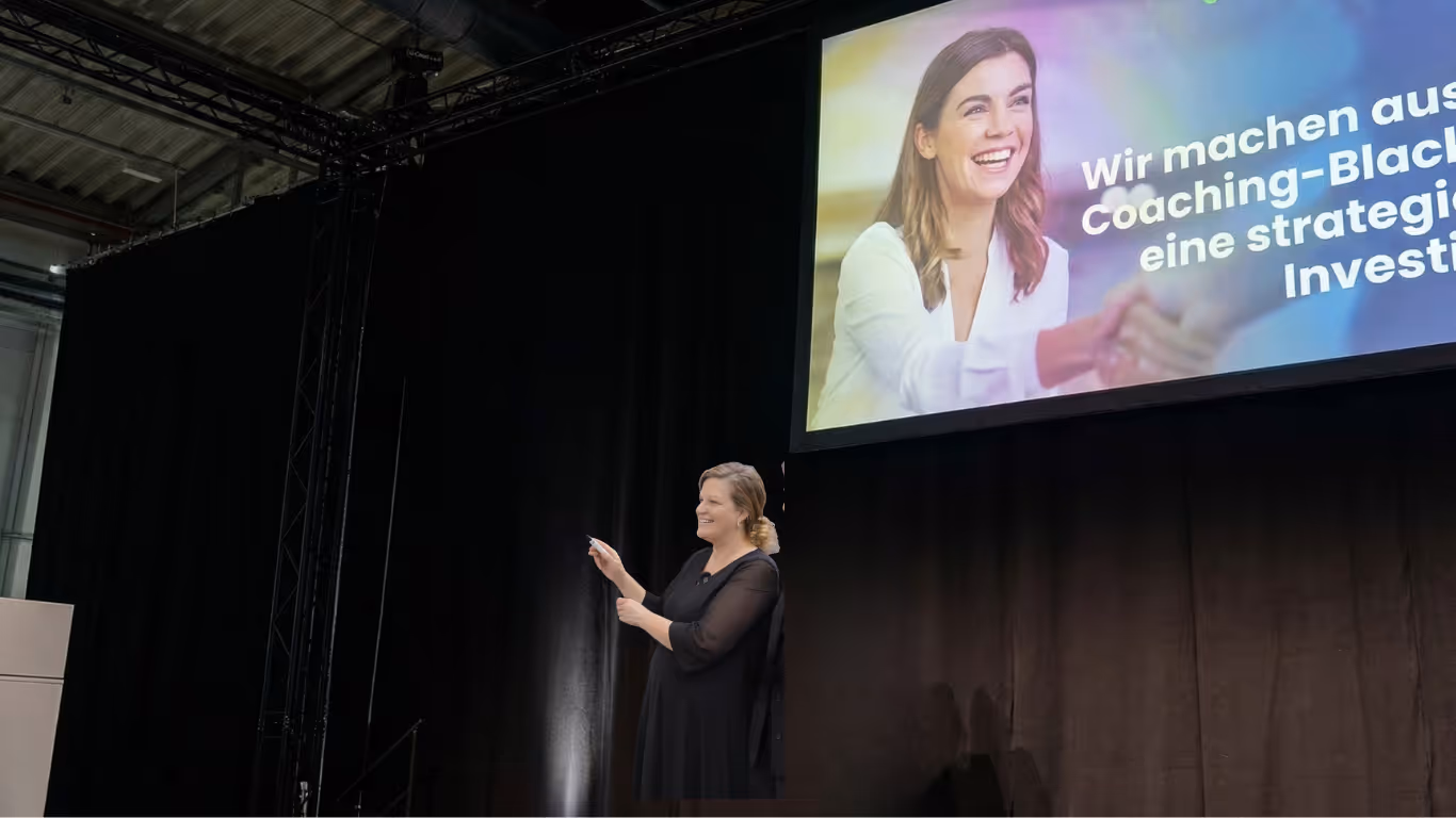 Nadia Argam in a black dress speaking and gesturing on stage with a large screen showing a smiling woman and partial German text in the background, pitching CoachMatcher to HR representatives.