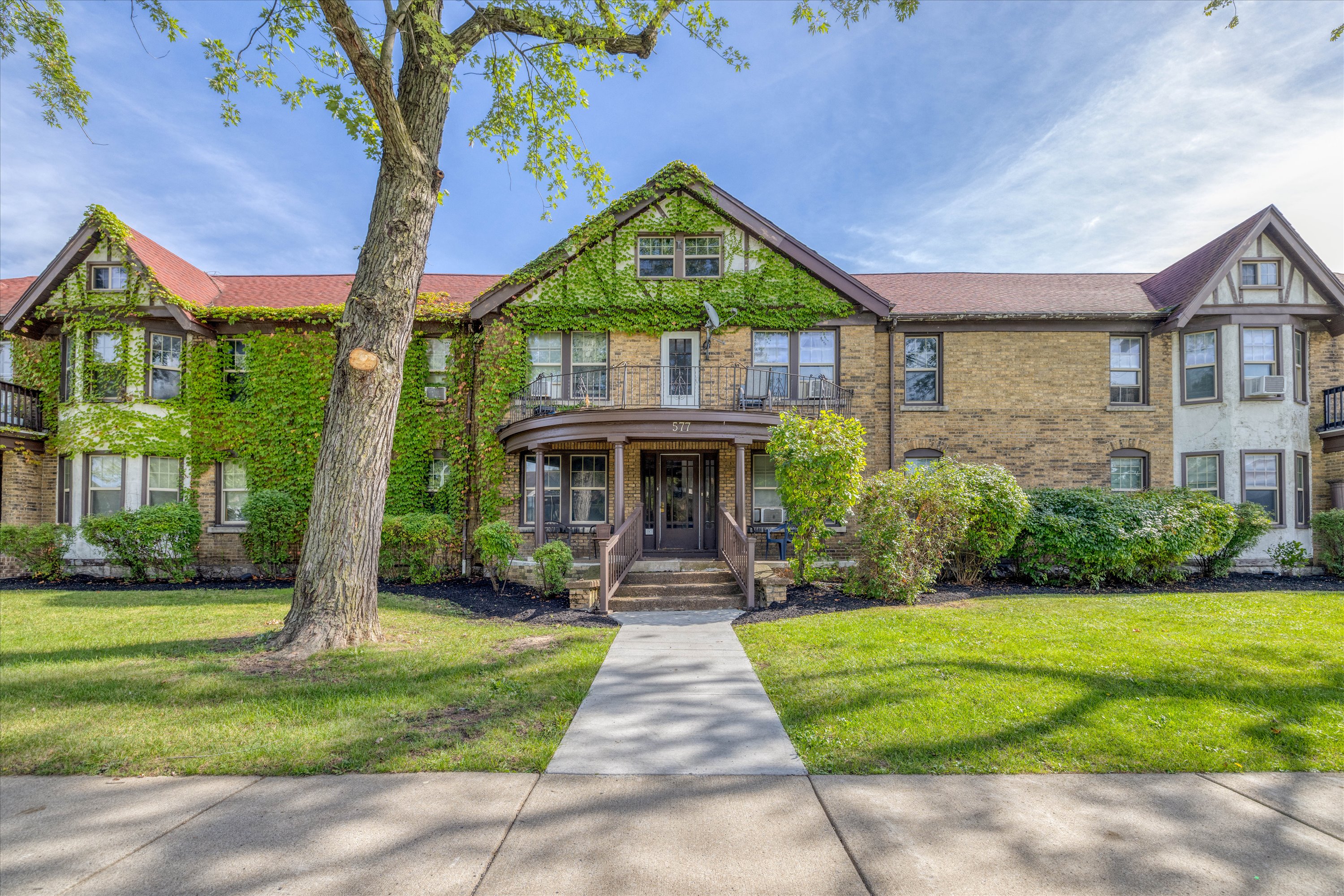 Two-story brick building with ivy growing on the facade, a central entrance with steps and a porch, surrounded by green lawns and shrubs under a blue sky.