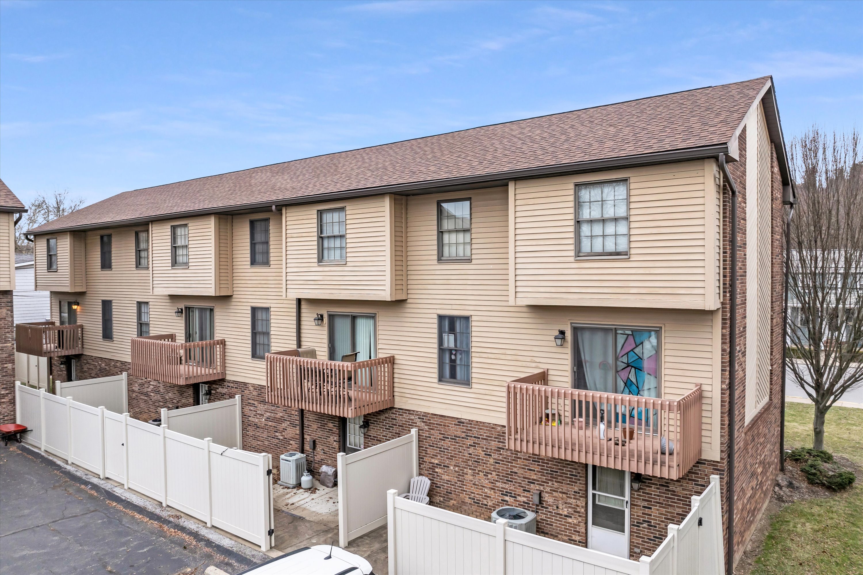 Two-story beige townhouse building with wooden balconies, brick lower walls, white fenced patios, and a parking area.
