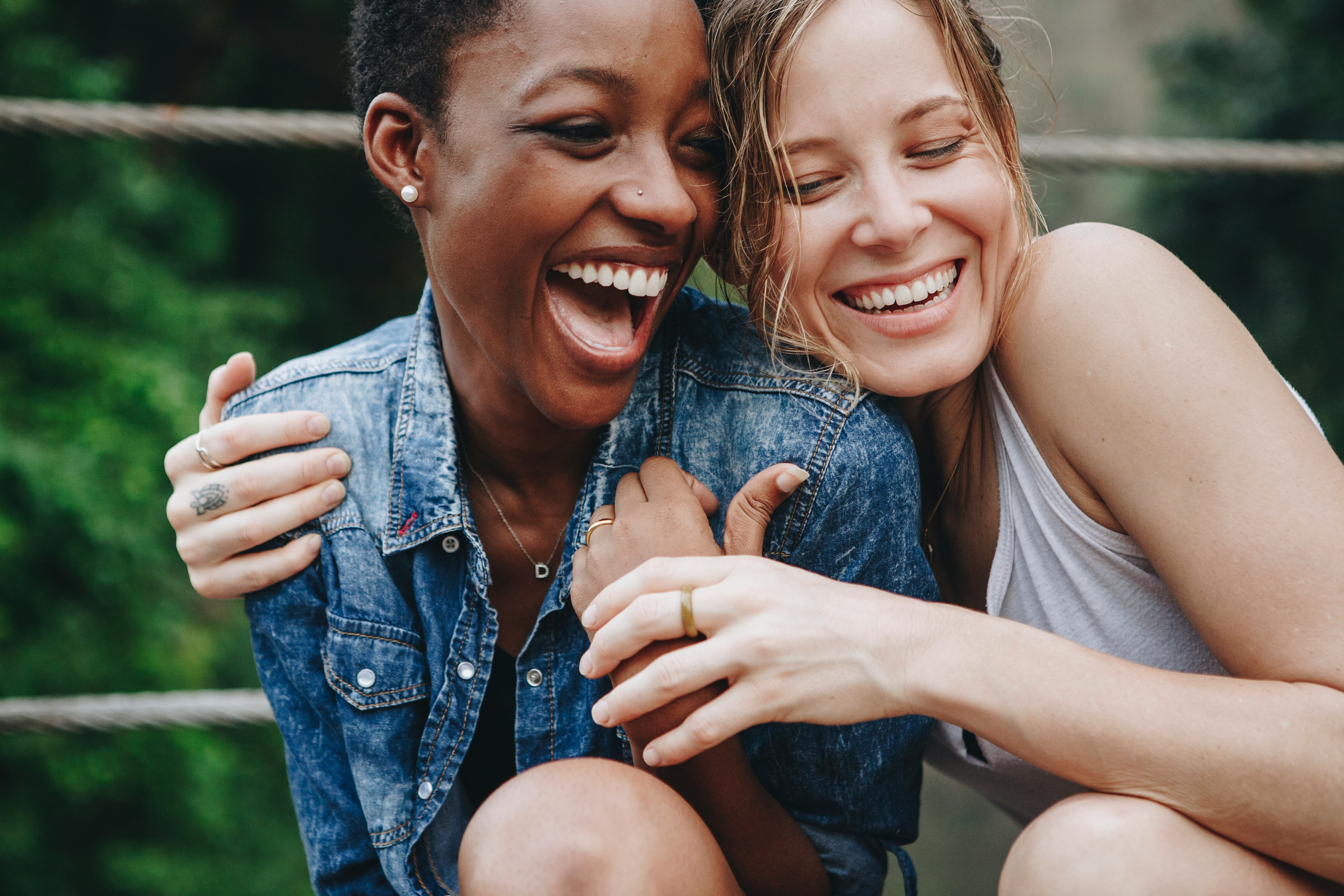 Two women laughing and hugging outdoors with greenery in the background.