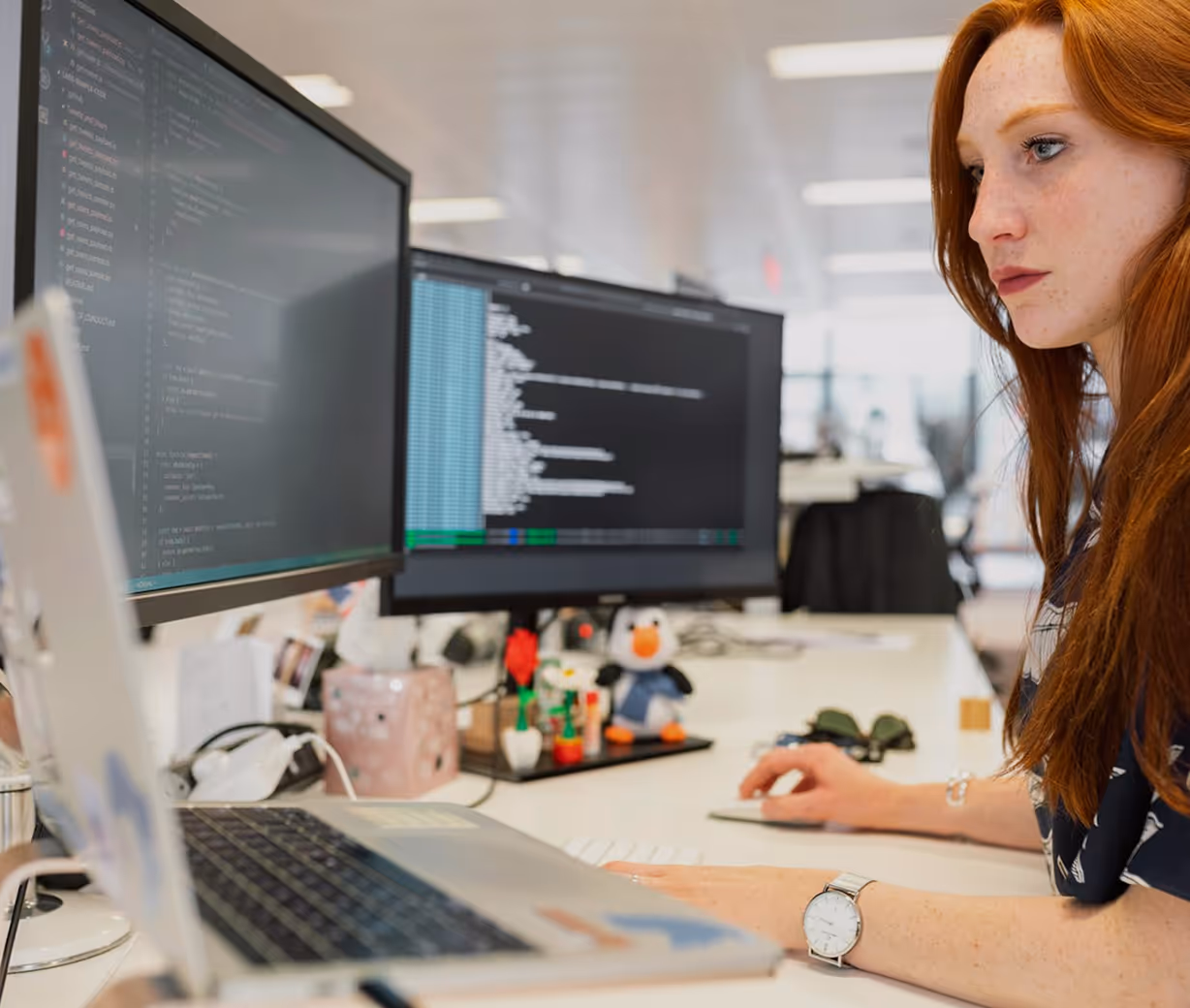 Red-haired woman focused on coding at her desk with dual monitors displaying programming code.