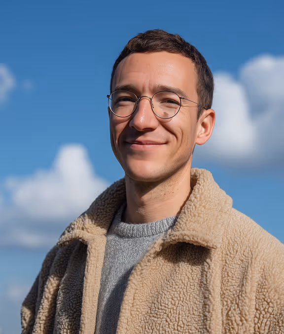 Smiling young man wearing round glasses and a beige fleece jacket against a blue sky with clouds.