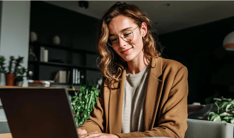 Woman wearing glasses and brown blazer using a laptop in a cozy room with plants and shelves.