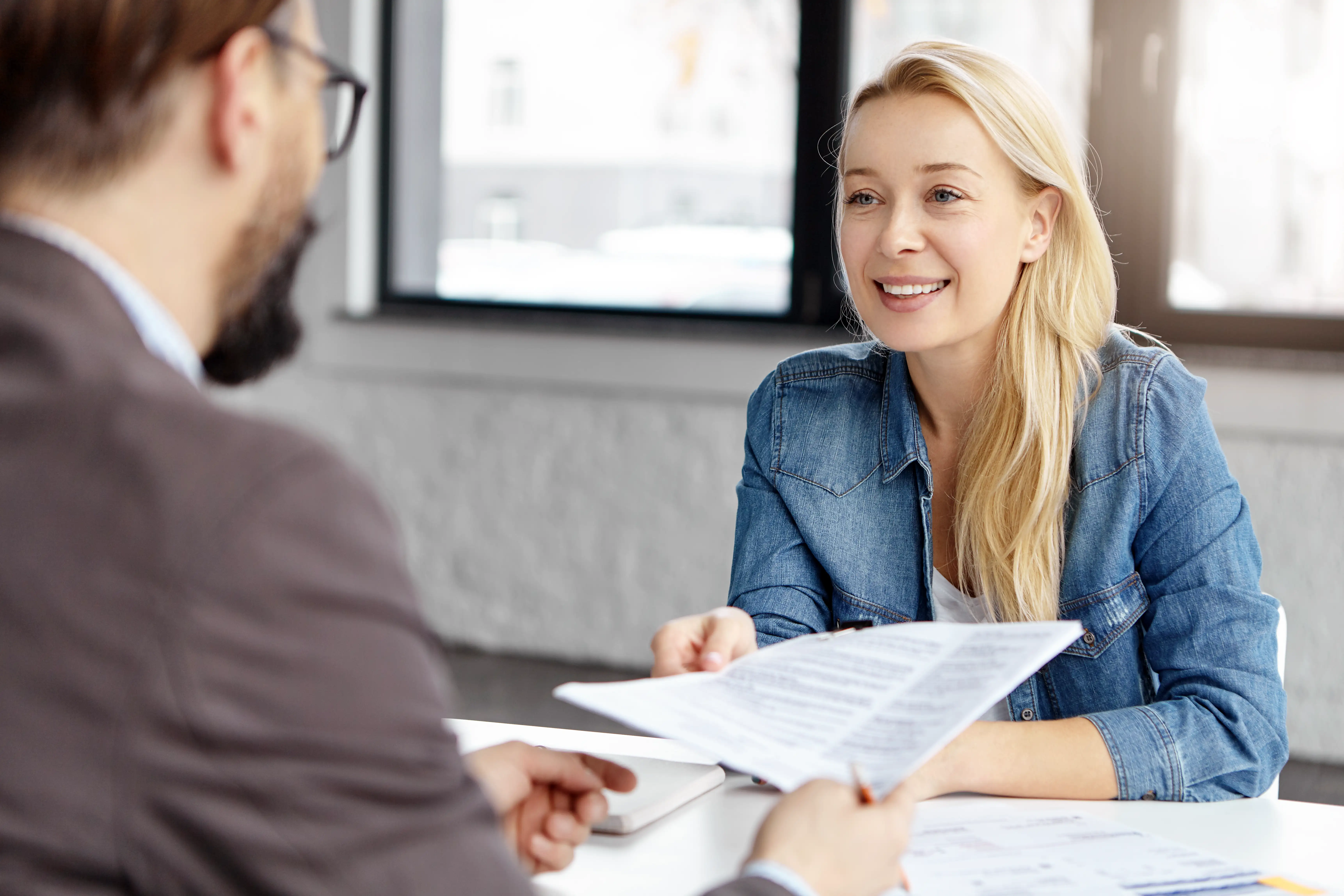Smiling woman handing documents to a man in glasses during a meeting at an office desk.