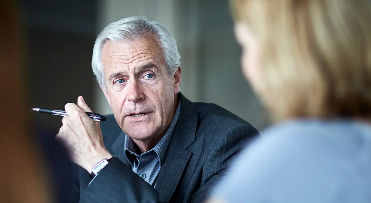 Older man in suit holding a pen, talking thoughtfully to two blurred people in a meeting.