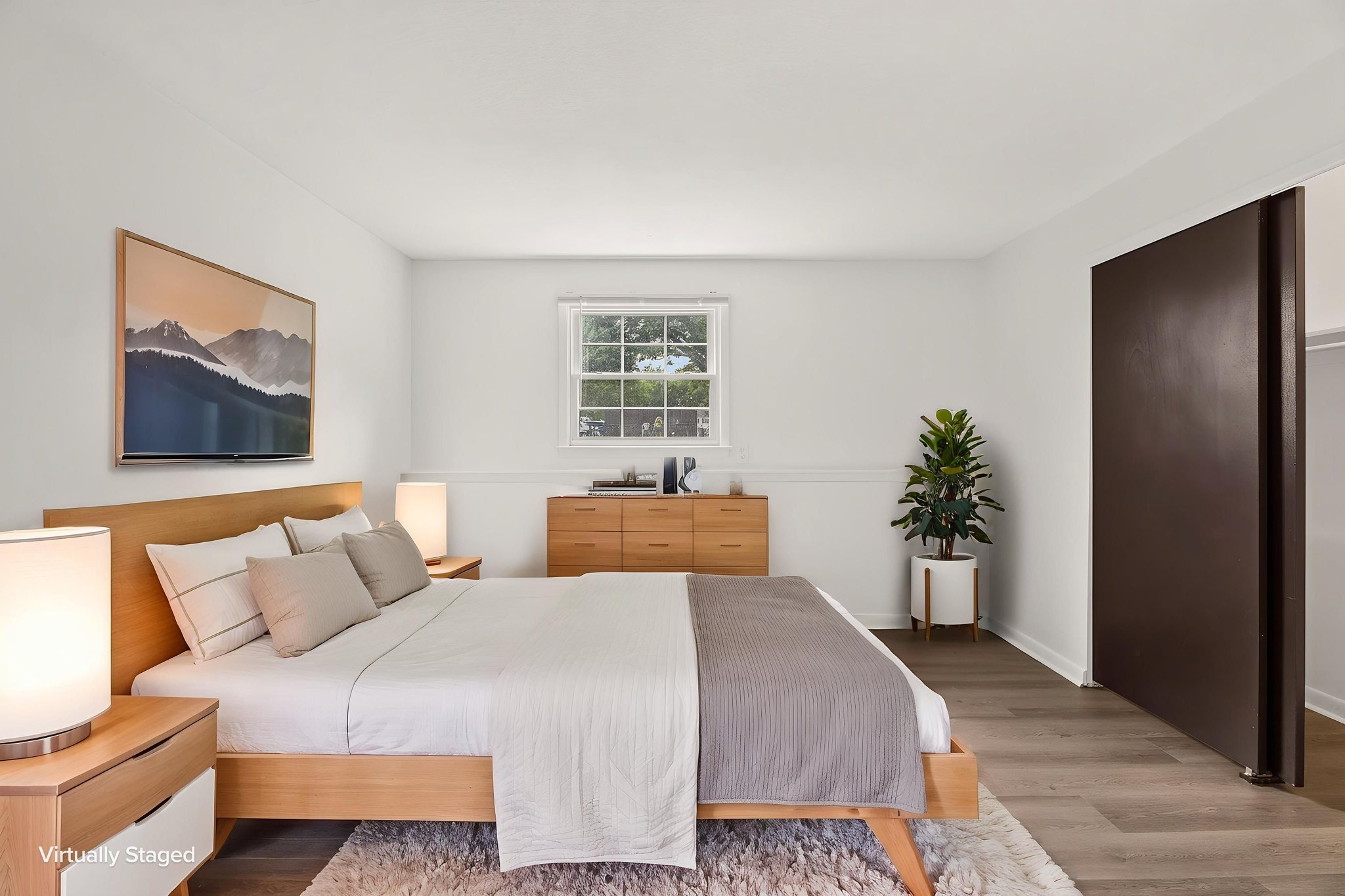 Virtually staged primary bedroom with a large bed, modern wood furniture, white walls, natural light, and spacious layout.
