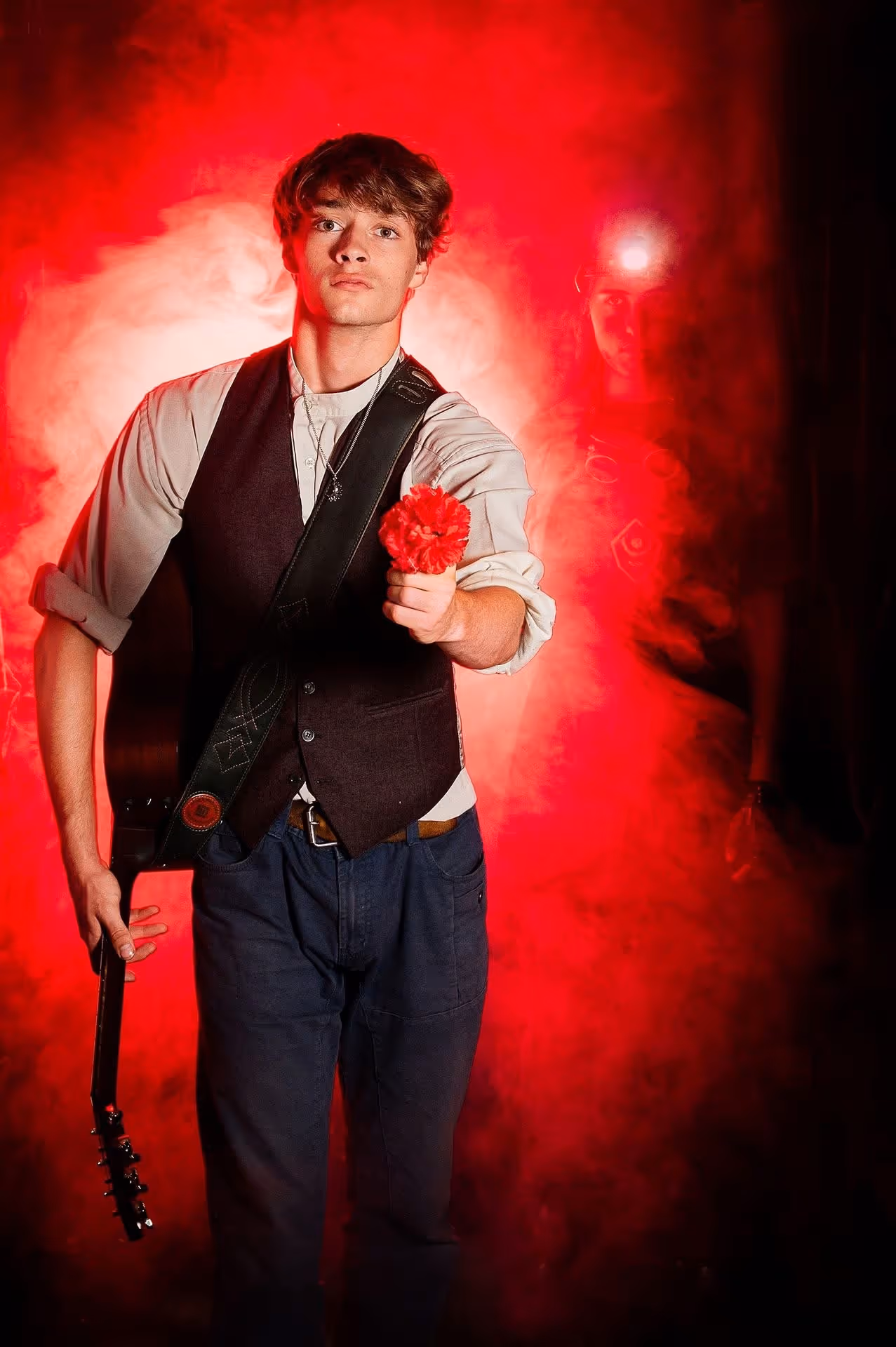 A lead performer stands facing forward, holding a red flower toward the camera and a guitar at their side. They wear a white shirt and dark vest, framed by red theatrical lighting and swirling smoke, with another performer faintly visible in the background.