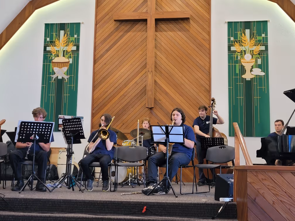 Students perform as a jazz ensemble onstage, seated with music stands and instruments including trombone, saxophone, drums, double bass, and piano. The group plays beneath tall wooden panelling, a large cross, and green hanging banners.