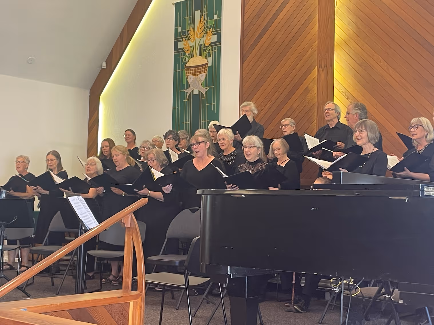 The choir performs during the Classical Finale Concert, singing from black choral folders onstage beside a grand piano, beneath warm wooden paneling and green hanging banners.